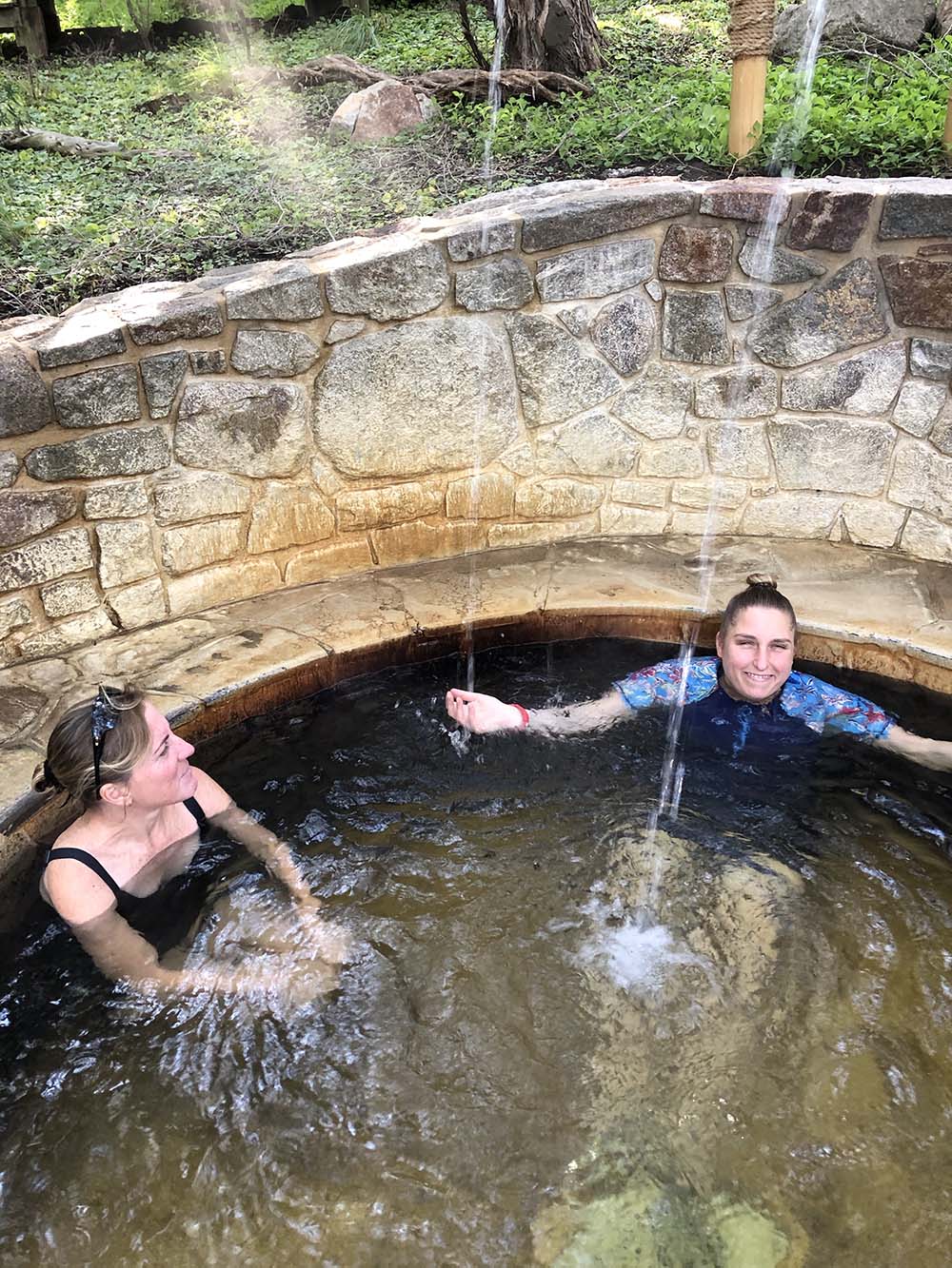 Two women sitting in stone bathing pool with small waterfall.